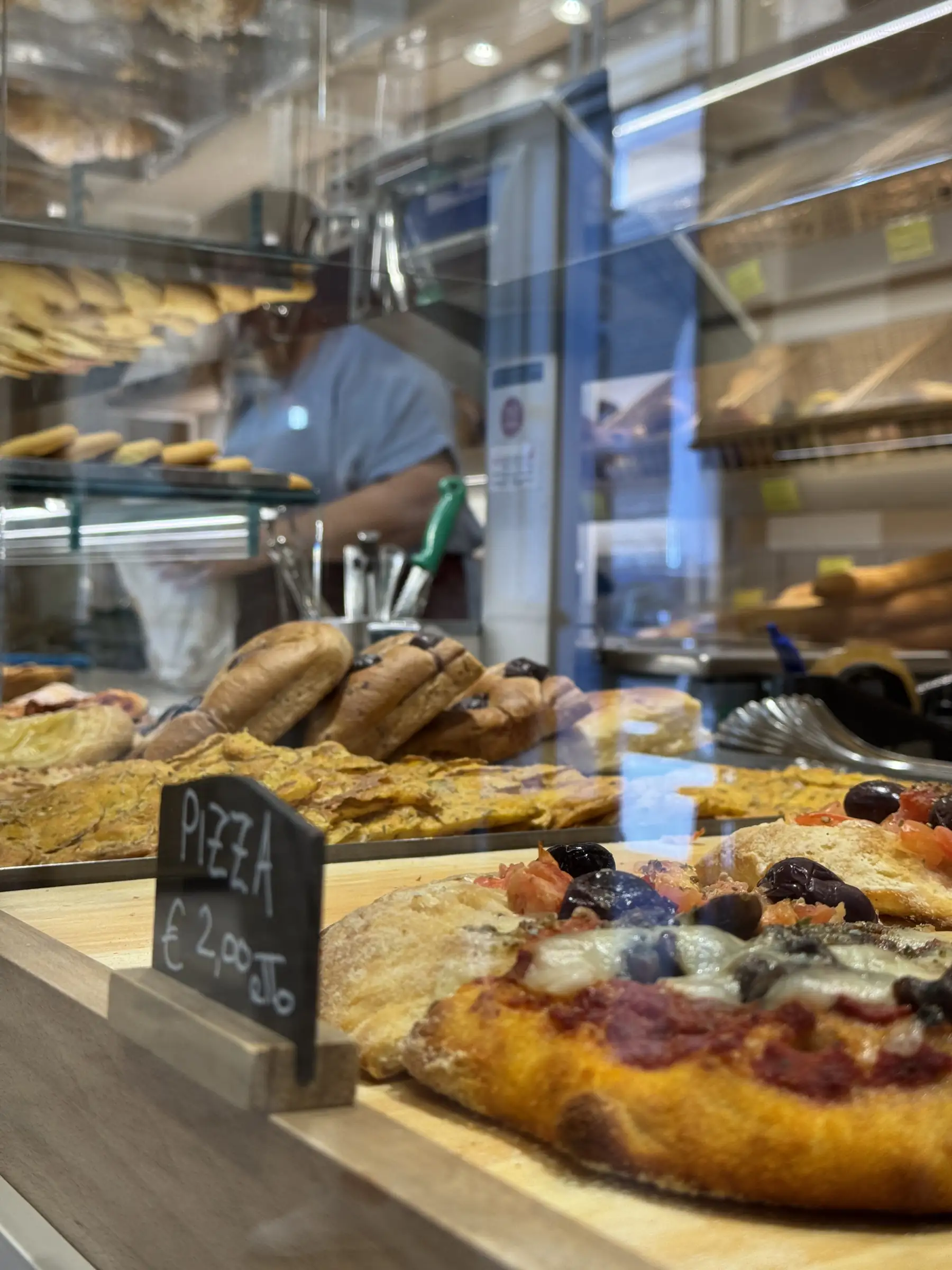 La vetrina del salato con le teglie di pizza e la lavagnetta, vista dal vetro del negozio